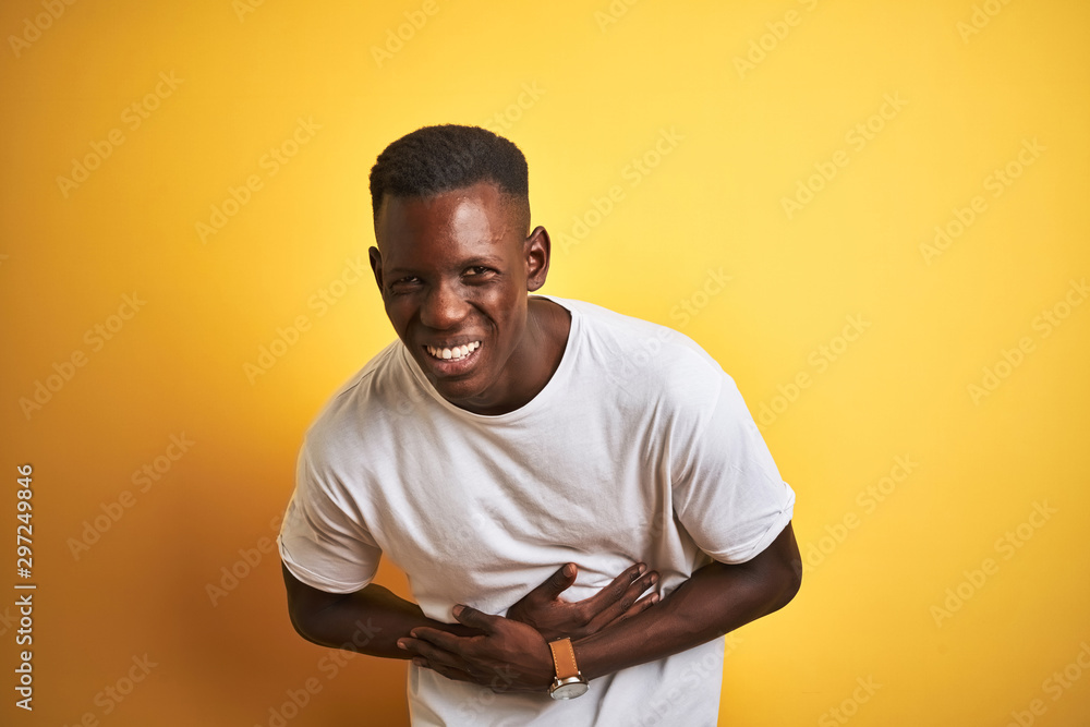 Young african american man wearing white t-shirt standing over isolated yellow background with hand on stomach because indigestion, painful illness feeling unwell. Ache concept. Young african american man wearing white t-shirt standing over isolated yellow background with hand on stomach because indigestion, painful illness feeling unwell. Ache concept.
