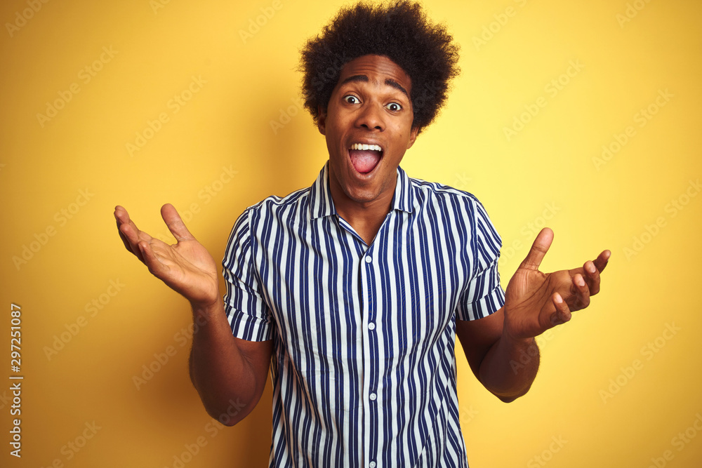 American man with afro hair wearing striped shirt standing over isolated yellow background celebrating crazy and amazed for success with arms raised and open eyes screaming excited. Winner concept American man with afro hair wearing striped shirt standing over isolated yellow background celebrating crazy and amazed for success with arms raised and open eyes screaming excited. Winner concept