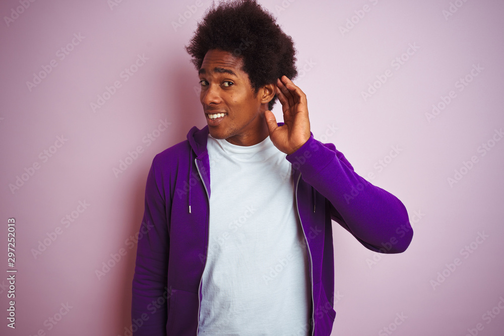 Young african american man wearing purple sweatshirt standing over isolated pink background smiling with hand over ear listening an hearing to rumor or gossip. Deafness concept. Young african american man wearing purple sweatshirt standing over isolated pink background smiling with hand over ear listening an hearing to rumor or gossip. Deafness concept.