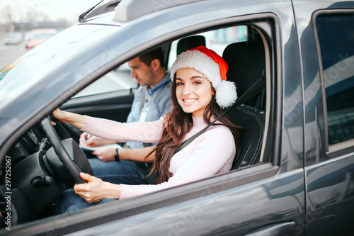 Young beautiful cheerful woman sit on driver's place in car. Holding hands on steering wheel and smile. Wear red hat. Christmas or new year period. Man sit besides her.