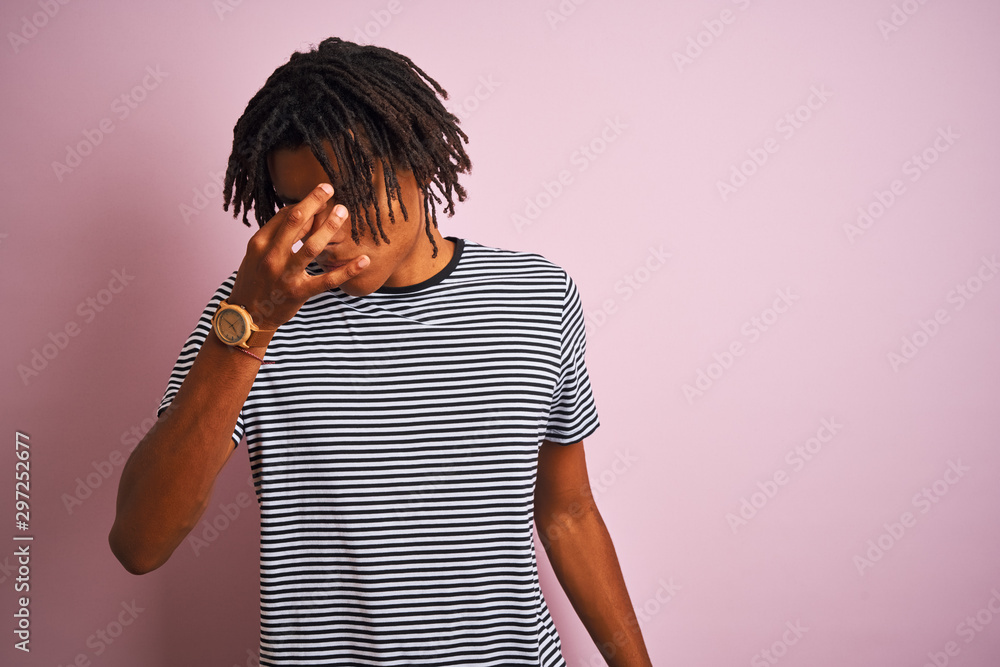 Afro man with dreadlocks wearing navy striped t-shirt standing over isolated pink background tired rubbing nose and eyes feeling fatigue and headache. Stress and frustration concept. Afro man with dreadlocks wearing navy striped t-shirt standing over isolated pink background tired rubbing nose and eyes feeling fatigue and headache. Stress and frustration concept.