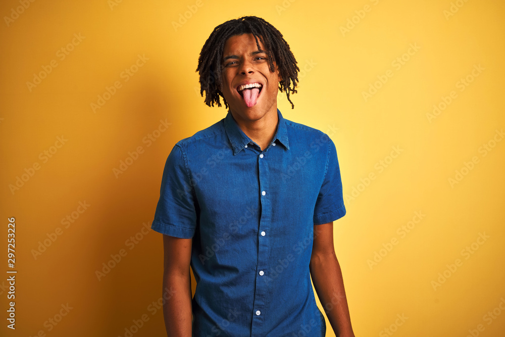 Afro man with dreadlocks wearing casual denim shirt standing over isolated yellow background sticking tongue out happy with funny expression. Emotion concept. Afro man with dreadlocks wearing casual denim shirt standing over isolated yellow background sticking tongue out happy with funny expression. Emotion concept.