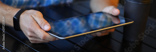 Focus on male hands holding high-tech tablet with blank display. Guy in classy outfit and fitness bracelet sitting at wooden table with notepad. Copy space on empty screen. Blurred background