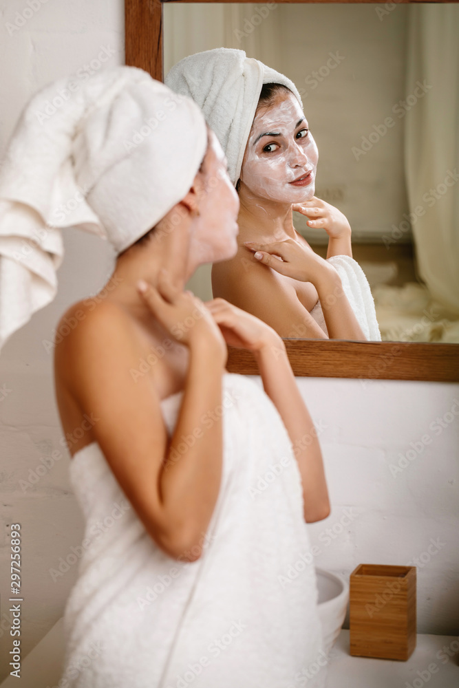 Woman with towel is applying mask on her face in front of the mirror at home. Skin care. Woman with towel is applying mask on her face in front of the mirror at home. Skin care.