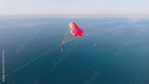 Towing a multicolored red parachute behind a boat jetski over the sea along the beach at sunset. Drone flied near the parachute. Aerial view.