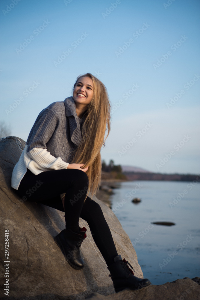 Young attractive woman sitting on a rock near the water. She's wearing a gray sweater and black pants. She smiles. Blue sky.