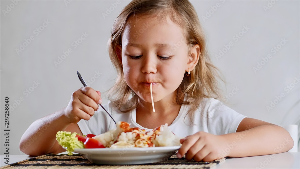 A little girl is eating spaghetti with vegetables at the table in the light room with a fork . Portrait of a kid eat at home in front of the camera. A little girl is eating spaghetti with vegetables at the table in the light room with a fork . Portrait of a kid eat at home in front of the camera.