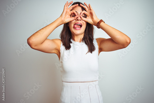 Young beautiful woman wearing dress standing over white isolated background doing ok gesture like binoculars sticking tongue out, eyes looking through fingers. Crazy expression.