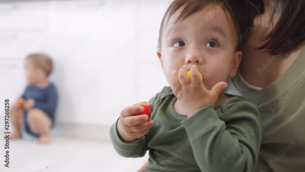 Medium shot of Asian baby sitting in mother’s lap, playing with wooden toy beads, putting one in mouth, dropping it and bending to pick up, and twin sibling in background Medium shot of Asian baby sitting in mother’s lap, playing with wooden toy beads, putting one in mouth, dropping it and bending to pick up, and twin sibling in background