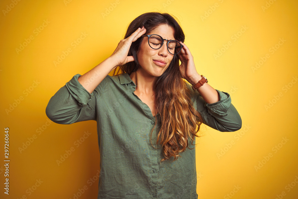 Young beautiful woman wearing green shirt and glasses over yelllow isolated background with hand on head for pain in head because stress. Suffering migraine. Young beautiful woman wearing green shirt and glasses over yelllow isolated background with hand on head for pain in head because stress. Suffering migraine.