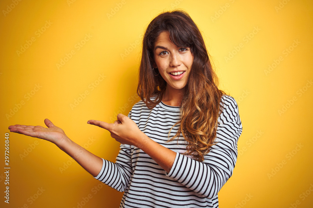 Young beautiful woman wearing stripes t-shirt standing over yelllow isolated background amazed and smiling to the camera while presenting with hand and pointing with finger. Young beautiful woman wearing stripes t-shirt standing over yelllow isolated background amazed and smiling to the camera while presenting with hand and pointing with finger.