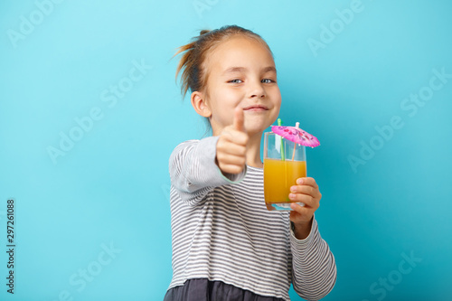 little child girl with orange juice, shows thumbs up, has a positive emotions, stands on blue backgound.