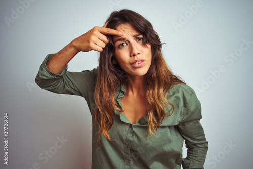 Young beautiful woman wearing green shirt standing over grey isolated background pointing unhappy to pimple on forehead, ugly infection of blackhead. Acne and skin problem