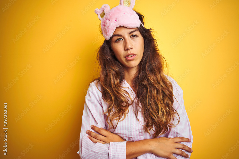 Naklejka premium Young woman wearing pajama and sleep mask standing over yellow isolated background looking sleepy and tired, exhausted for fatigue and hangover, lazy eyes in the morning.