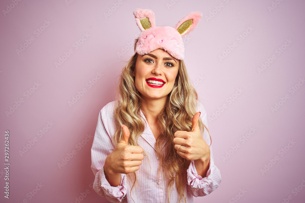 Young beautiful woman wearing pajama and sleep mask over pink isolated background success sign doing positive gesture with hand, thumbs up smiling and happy. Cheerful expression and winner gesture. Young beautiful woman wearing pajama and sleep mask over pink isolated background success sign doing positive gesture with hand, thumbs up smiling and happy. Cheerful expression and winner gesture.