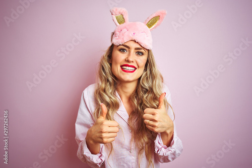 Young beautiful woman wearing pajama and sleep mask over pink isolated background success sign doing positive gesture with hand, thumbs up smiling and happy. Cheerful expression and winner gesture.