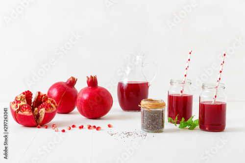 Pitcher and two jars of fresh pomegranate juice, with three ripe pomegranates, one freshly peeled. Rouge pomegranate seeds and chia seeds in the foreground, all on a white table. photo