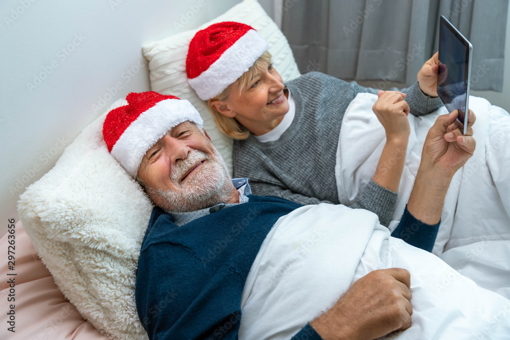 Obraz premium Senior couple using tablet to video phone call to greet their family aboard in different time zone for Christmas festival on bed in bedroom; looking at camera