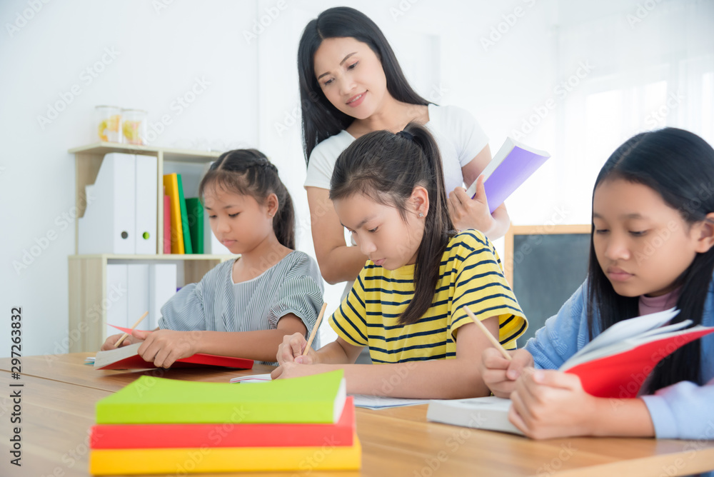 Three asian students sitting and reading book in the classroom with a teacher standing by. Three asian students sitting and reading book in the classroom with a teacher standing by.