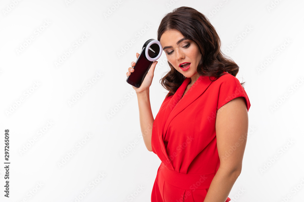 Obraz premium A young woman with red lips, bright makeup, dark wavy long hair, in a red suit, black glasses with transparent stacks stands and holds the phone in her hands