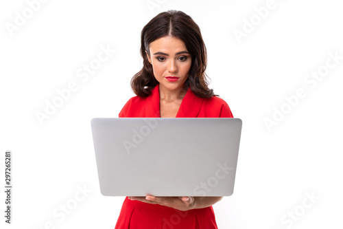 A young woman with red lips, bright makeup, dark wavy long hair, in a red suit, stands with a white laptop