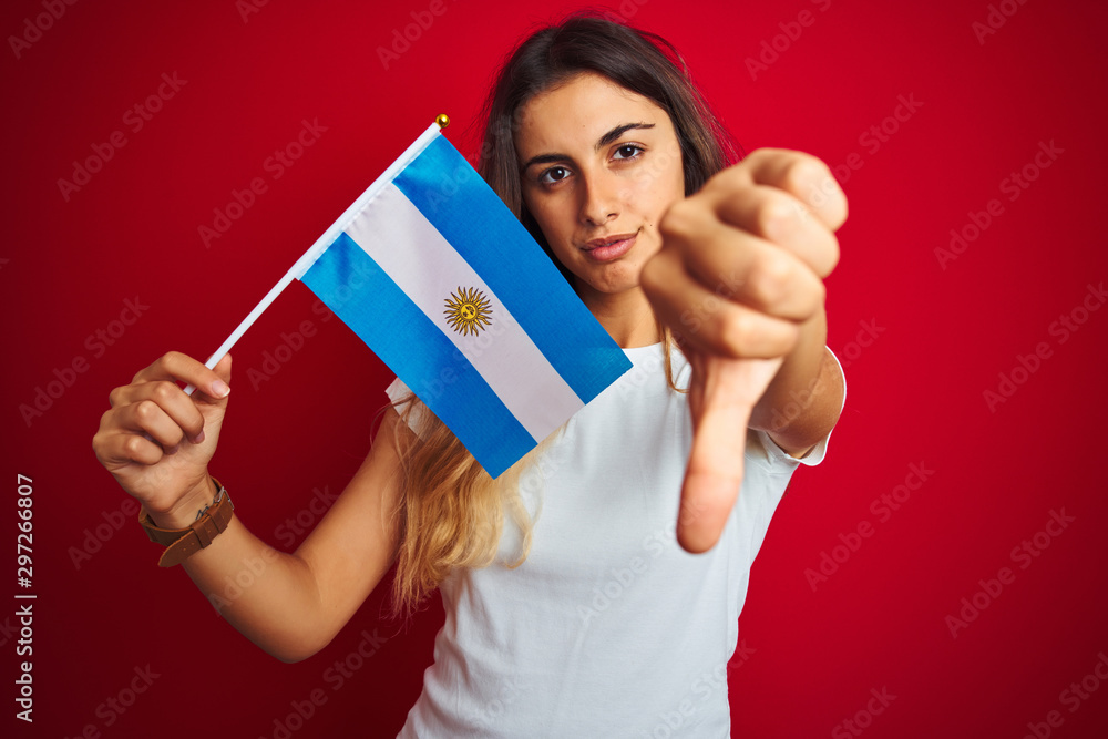 Young beautiful woman holding argentine flag over red isolated background with angry face, negative sign showing dislike with thumbs down, rejection concept Young beautiful woman holding argentine flag over red isolated background with angry face, negative sign showing dislike with thumbs down, rejection concept
