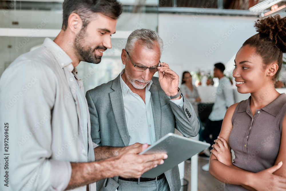 Let me see. Confident mature business man in formal wear looking at digital tablet while standing with young coworkers in the modern office Let me see. Confident mature business man in formal wear looking at digital tablet while standing with young coworkers in the modern office