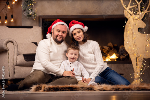 Photo of man and woman in Santa s cap with son sitting on floor on background of fireplace