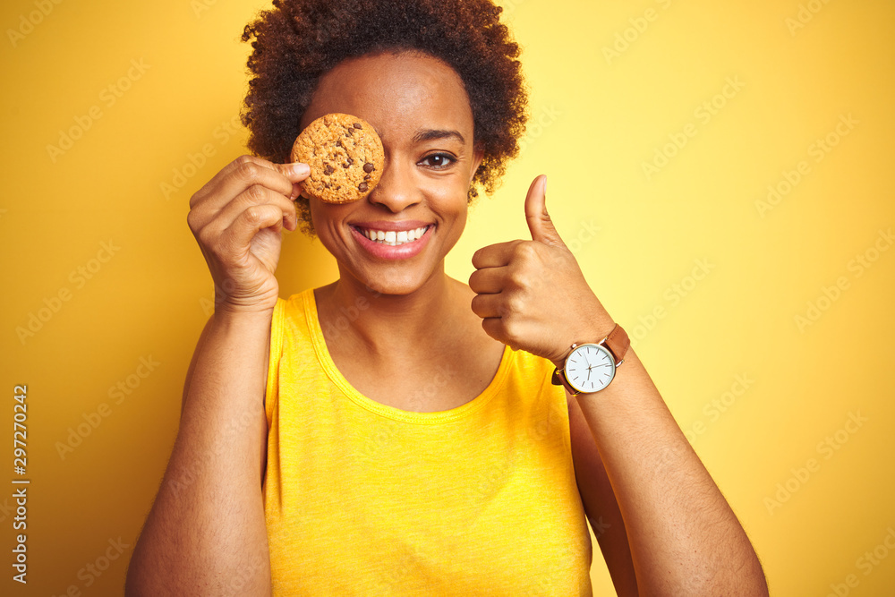 African american woman holding chocolate chips cookie over yellow background happy with big smile doing ok sign, thumb up with fingers, excellent sign African american woman holding chocolate chips cookie over yellow background happy with big smile doing ok sign, thumb up with fingers, excellent sign