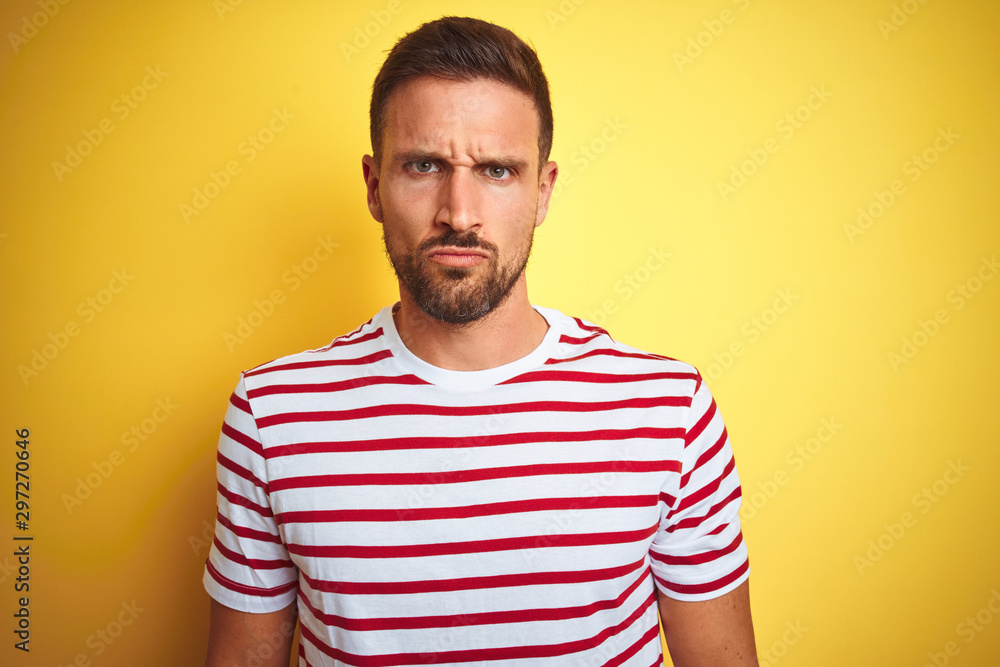 Young handsome man wearing casual red striped t-shirt over yellow isolated background skeptic and nervous, frowning upset because of problem. Negative person. Young handsome man wearing casual red striped t-shirt over yellow isolated background skeptic and nervous, frowning upset because of problem. Negative person.