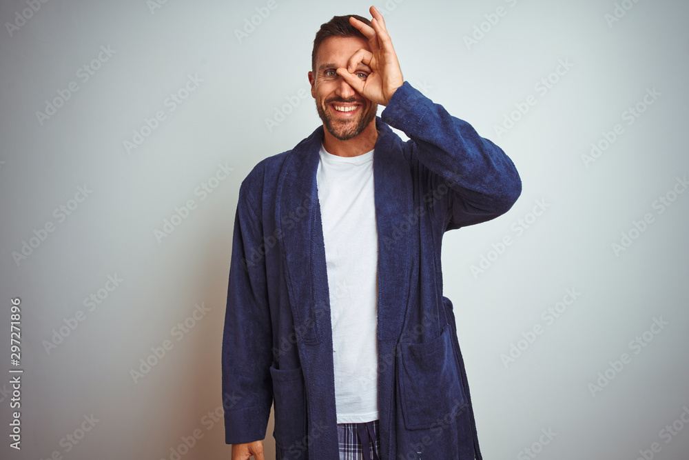 Young man wearing comfortable pajamas and robe at morning over isolated background doing ok gesture with hand smiling, eye looking through fingers with happy face. Young man wearing comfortable pajamas and robe at morning over isolated background doing ok gesture with hand smiling, eye looking through fingers with happy face.
