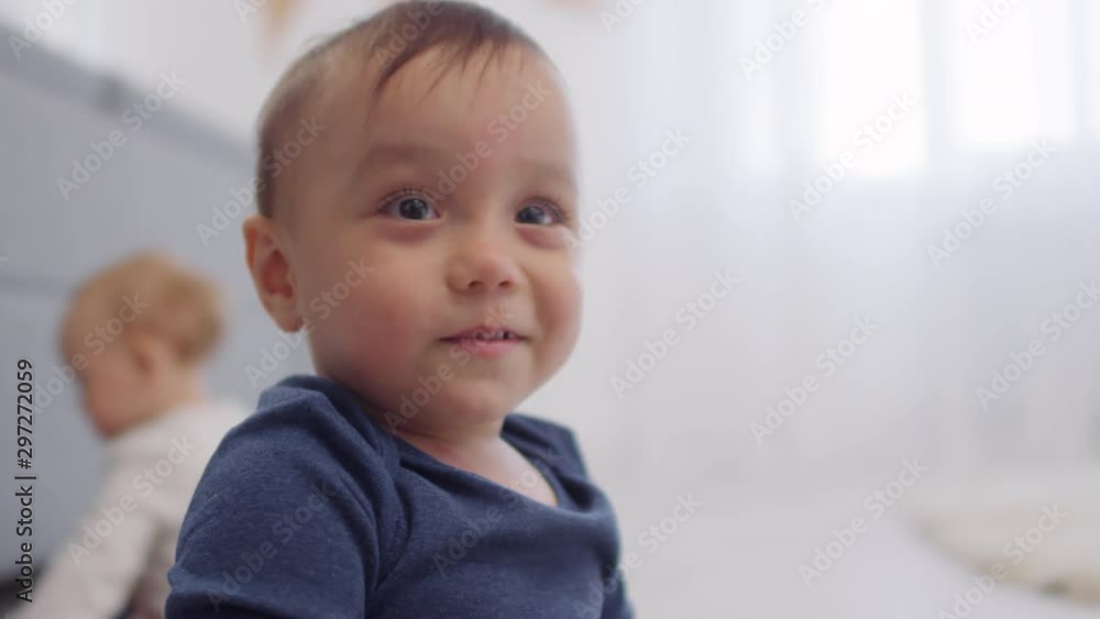 Close-up shot of Asian baby looking at something towards camera then smiling happily, and twin sibling sitting and playing in background Close-up shot of Asian baby looking at something towards camera then smiling happily, and twin sibling sitting and playing in background