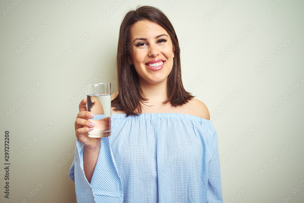 Young beautiful woman drinking a glass of fresh water over isolated background with a happy face standing and smiling with a confident smile showing teeth Young beautiful woman drinking a glass of fresh water over isolated background with a happy face standing and smiling with a confident smile showing teeth