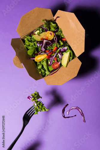 Mixed salad in cardbox on purple background with fork. Healthcare concept. Top view.