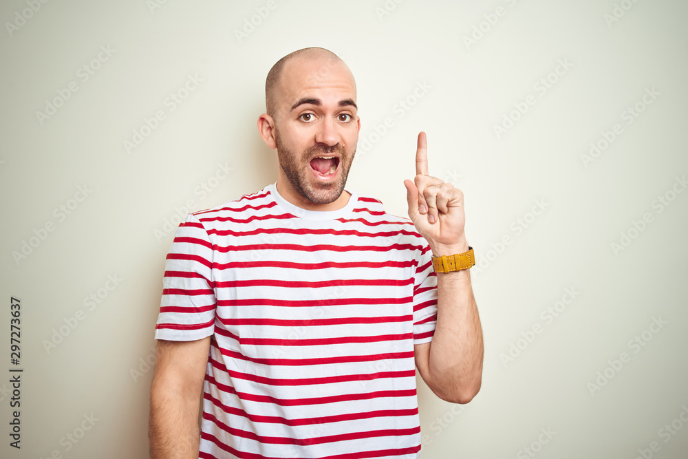 Naklejka premium Young bald man with beard wearing casual striped red t-shirt over white isolated background pointing finger up with successful idea. Exited and happy. Number one.