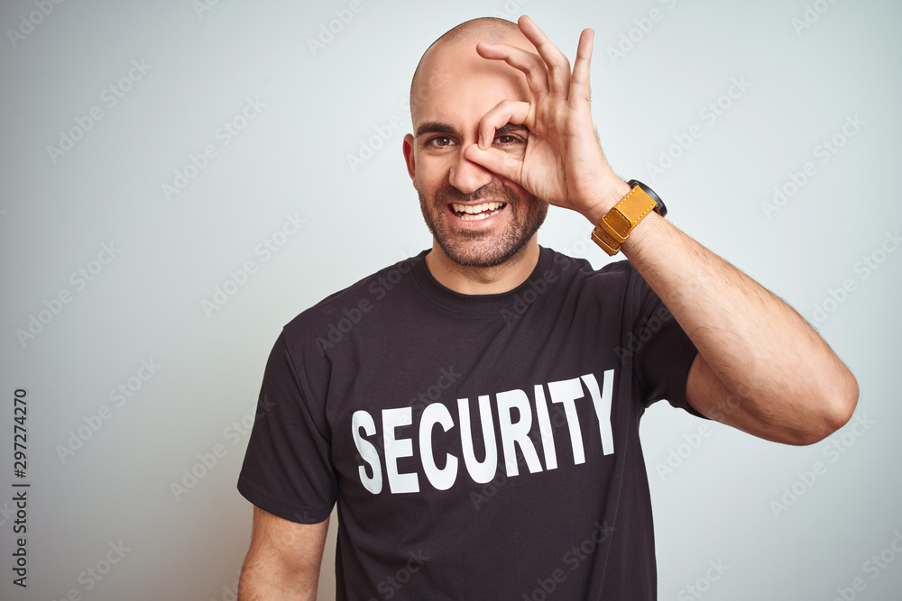 Young safeguard man wearing security uniform over isolated background doing ok gesture with hand smiling, eye looking through fingers with happy face. - obrazy, fototapety, plakaty 