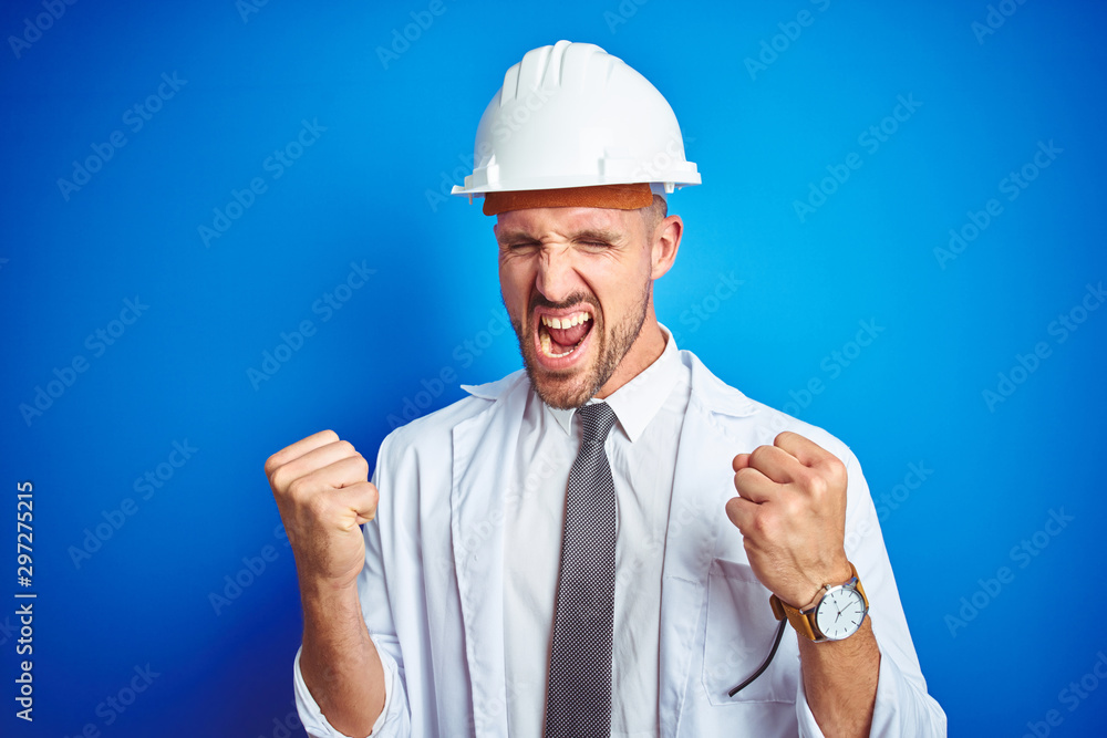 Young handsome engineer man wearing safety helmet over blue isolated background very happy and excited doing winner gesture with arms raised, smiling and screaming for success. Celebration concept.