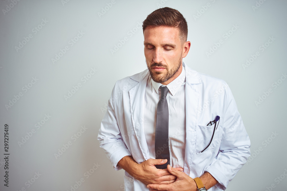 Young handsome doctor man wearing white profressional coat over isolated background with hand on stomach because nausea, painful disease feeling unwell. Ache concept. Young handsome doctor man wearing white profressional coat over isolated background with hand on stomach because nausea, painful disease feeling unwell. Ache concept.