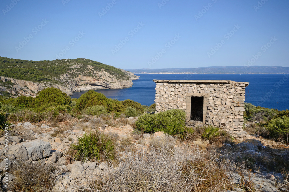 the highest point of Punta Giglio in the Mediterranean coast of Sardinia. In the foreground the remains of a fascist war post from World War II and in the background the city of Alghero