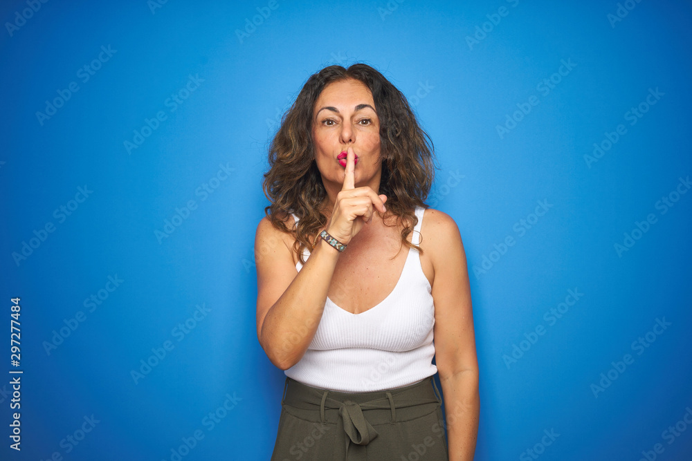 Middle age senior woman with curly hair standing over blue isolated background asking to be quiet with finger on lips. Silence and secret concept.
