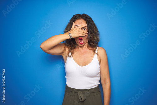 Middle age senior woman with curly hair standing over blue isolated background peeking in shock covering face and eyes with hand, looking through fingers with embarrassed expression. © Krakenimages.com