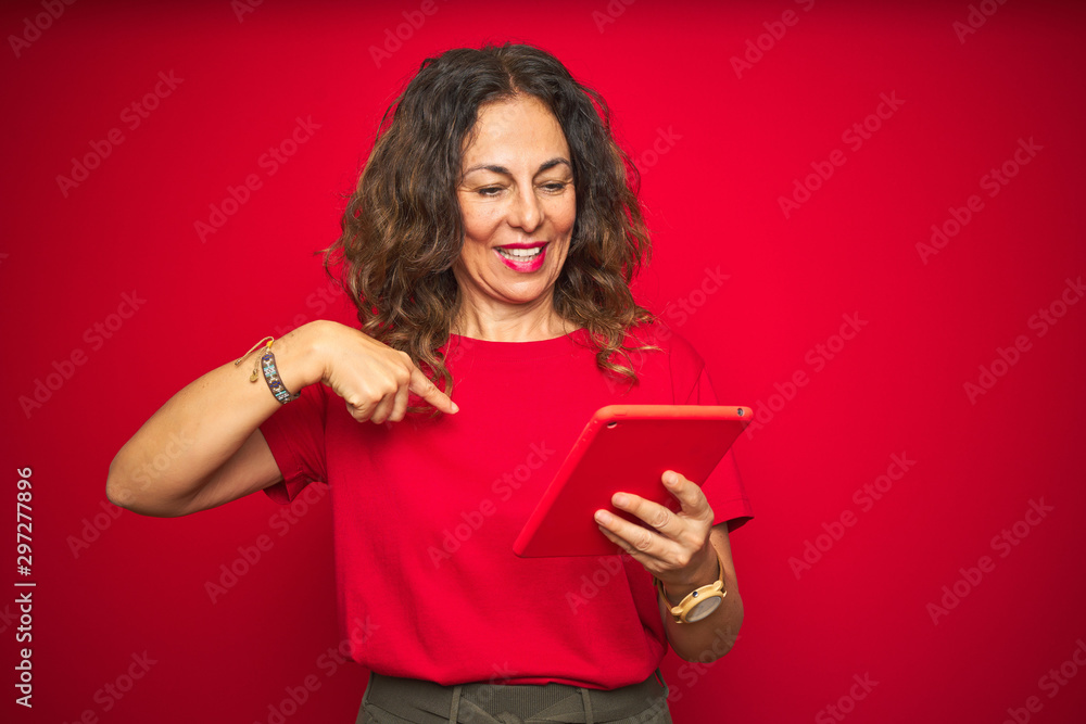 Middle age senior woman using touchpad tablet over red isolated background with surprise face pointing finger to himself Middle age senior woman using touchpad tablet over red isolated background with surprise face pointing finger to himself