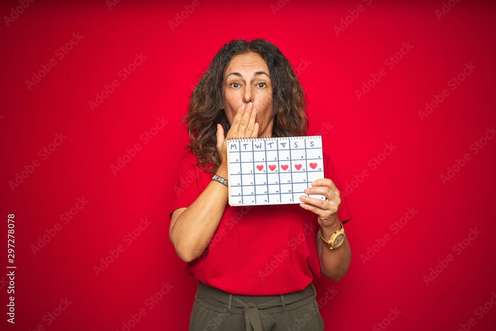 Middle age senior woman holding menstruation calendar over red isolated background cover mouth with hand shocked with shame for mistake, expression of fear, scared in silence, secret concept Middle age senior woman holding menstruation calendar over red isolated background cover mouth with hand shocked with shame for mistake, expression of fear, scared in silence, secret concept