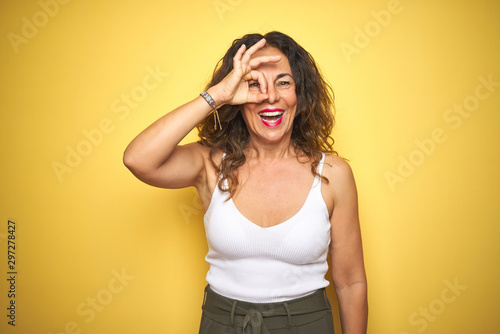 Middle age senior woman with curly hair standing over yellow isolated background doing ok gesture with hand smiling, eye looking through fingers with happy face.