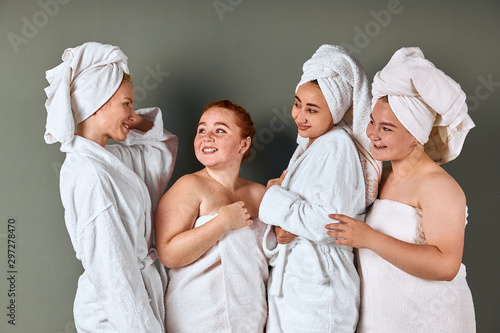 Beautiful young smiling women with perfect skin wearing white bathing towels having fun isolated on grey background