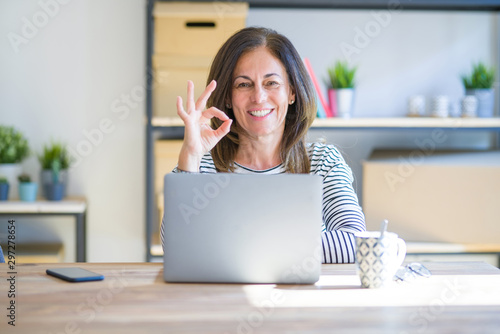 Foto Middle age senior woman sitting at the table at home working using computer laptop smiling positive doing ok sign with hand and fingers