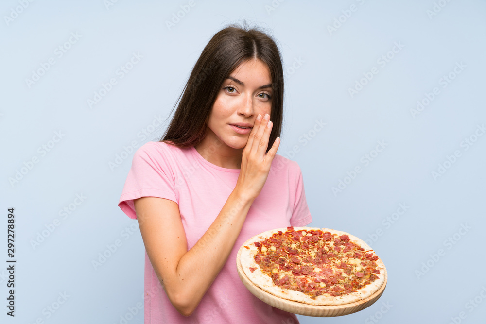 Pretty young girl holding a pizza over isolated blue wall whispering something
