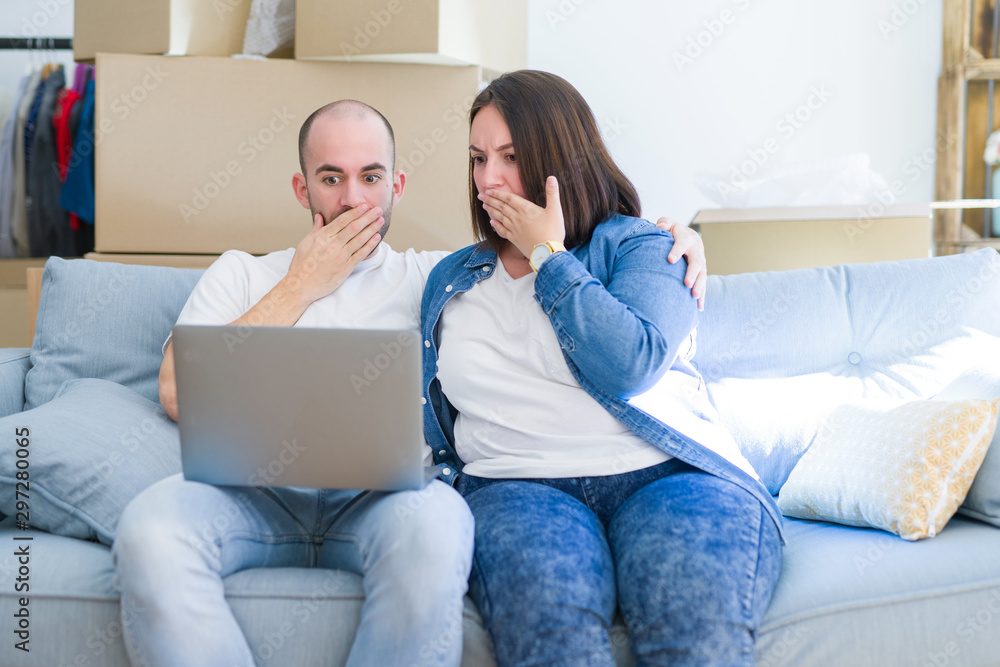 Young couple sitting on the sofa arround cardboard boxes moving to a new house using laptop cover mouth with hand shocked with shame for mistake, expression of fear, scared in silence, secret concept Young couple sitting on the sofa arround cardboard boxes moving to a new house using laptop cover mouth with hand shocked with shame for mistake, expression of fear, scared in silence, secret concept