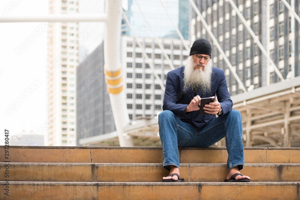 Mature bearded businessman sitting while using phone in the city outdoors Mature bearded businessman sitting while using phone in the city outdoors