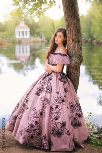 Fototapeta A beautiful young woman in a 19th-century dress stands with a book in a park